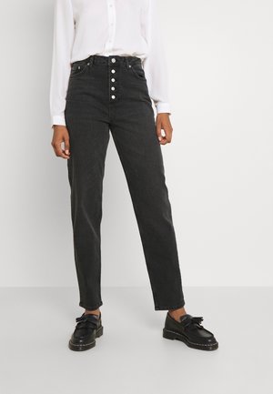 Person iført sorte højtaljede jeans med forreste knapper, hvid langærmet skjorte og sorte læder loafers med dusk på en hvid baggrund.