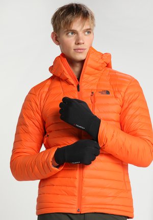 Bright orange puffer jacket with a high collar, horizontal quilted pattern, and a zippered pocket. Black gloves with Polartec branding.