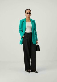 Türkiser Blazer über einem weißen Oberteil, kombiniert mit weit geschnittenen schwarzen Hosen. Die Accessoires umfassen eine kleine schwarze Handtasche und dunkle Sonnenbrillen.