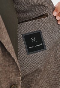 Brązowy materiał w wzory z subtelną teksturą. Zawiera czarną metkę z białymi detalami logo, wszytą wewnątrz odzieży.