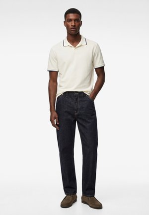 Man staat met één hand in de zak, draagt een crème korte mouwen polo, donkere jeans en bruine schoenen tegen een witte achtergrond.