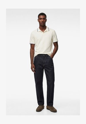 Uomo in piedi con una mano in tasca che indossa una polo color crema a maniche corte, jeans scuri e scarpe marroni su sfondo bianco.