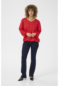 Rotes Langarm-T-Shirt mit V-Ausschnitt, lockerer Passform, gerafften Ärmelabschlüssen, kombiniert mit dunkelblauen, enganliegenden Jeans und schwarzen, spitz zulaufenden Absätzen.