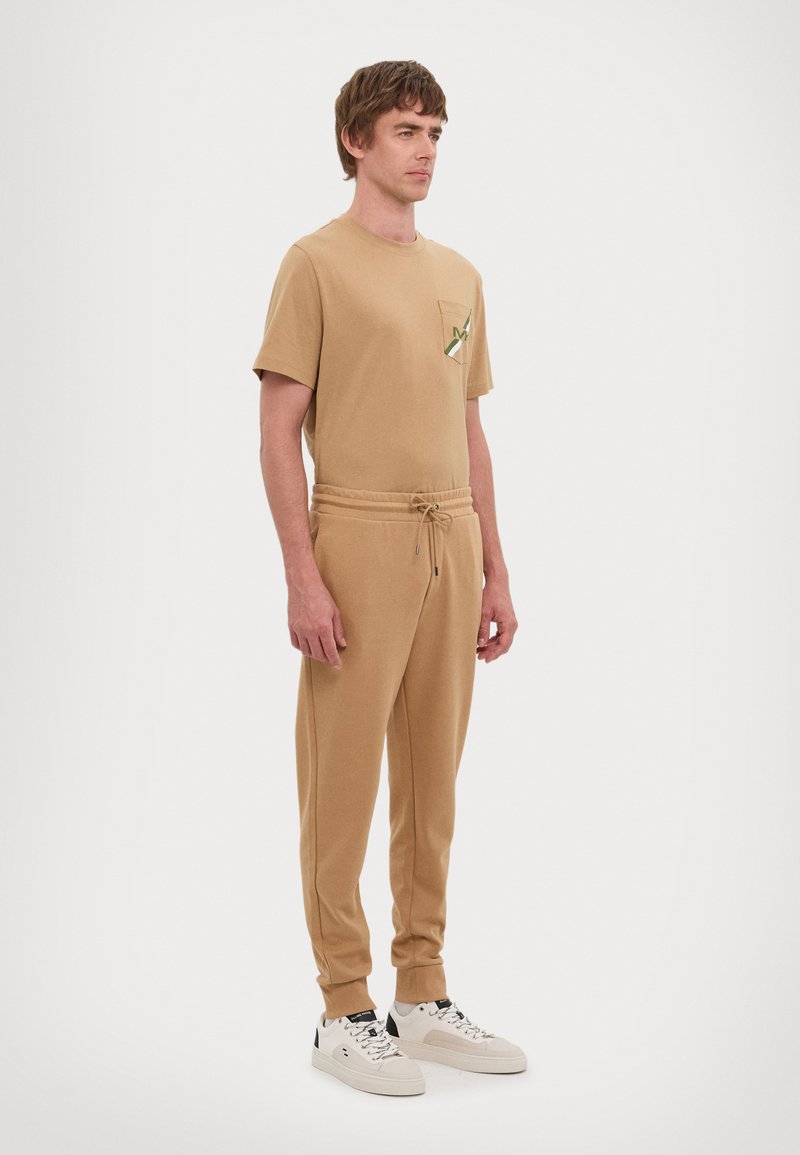 Camiseta de algodón beige con un bolsillo y un diseño verde, combinada con pantalones de tiro ajustable a juego. El modelo lleva zapatillas blancas con acentos negros.