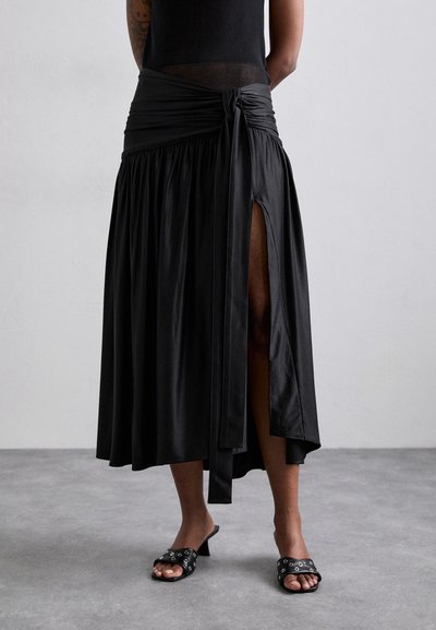 Μαύρη σατινένια φούστα με σ gathered waist, πλαϊνό σκίσιμο και κόμπο στη λεπτομέρεια; συνδυασμένη με μαύρες μπαλαρίνες με καρφίτσες.