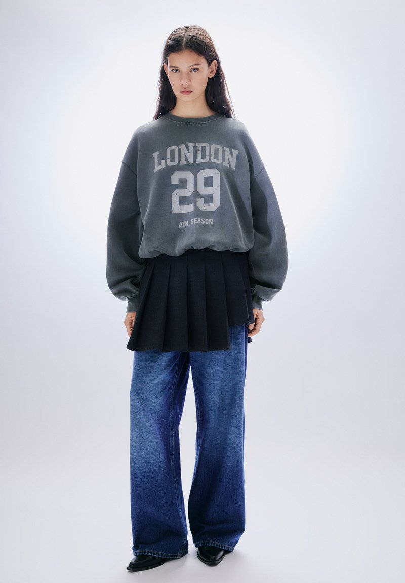 Harmaa collegepaita, jossa valkoisella painettu teksti "LONDON 29", yhdistetty mustaan pliseerattuun hameeseen ja tummansinisiin leveälahkeisiin farkkuihin. Mustat teräväkärkiset kengät.