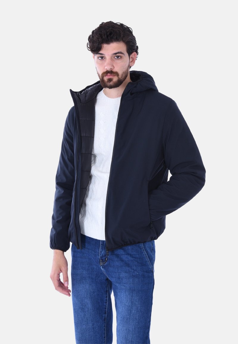 Giubbotto piumino blu navy con cappuccio staccabile, pannello frontale trapuntato e polsini elastici. Abbinato a un maglione bianco a treccia e jeans blu.