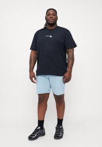 Svart t-skjorte med korte ermer og hvit tekst, kombinert med lyseblå denimshorts. Iført svarte treningsko og svarte sokker.