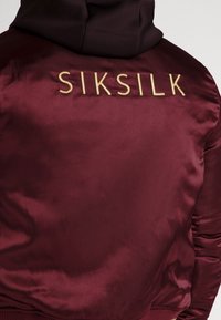 Burgunder satinvindjakke med hætte, prydet med guld "SIKSILK" tekst på bagsiden, ribbede manchetter og en glat tekstur.
