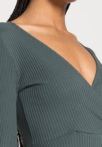 Kvinna iförd en ribbad, djup V-ringad, långärmad mörkgrön topp som visar dekolettage och flätat hår över axeln.