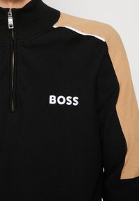 Pull noir à fermeture éclair quart de zip avec manches beige et texte blanc "BOSS" sur la poitrine, avec un col montant et un curseur de zip.