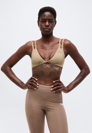 Urheilullinen nainen lyhyissä hiuksissa, beige urheiluliivi ja leggingsit päällä, seisoo käsivarret lanteilla yksiväristä taustaa vasten.