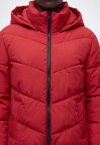 Personne portant une veste matelassée rouge avec une capuche et une fermeture éclair centrale noire, montrant la partie supérieure du torse et la zone du cou.