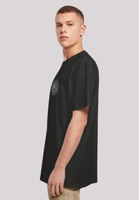 Schwarzes Baumwoll-T-Shirt mit lockerer Passform, mit einem kreisförmigen grafischen Design in Grau auf der linken Brust. Kurze Ärmel und runder Halsausschnitt.