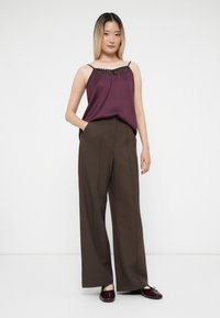 Camisola in seta bordeaux con scollatura tempestata di paillettes, abbinata a pantaloni larghi marroni. Il modello mostra una tasca frontale e scarpe bordeaux piatte e lucide.