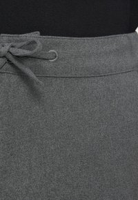 Prim-plan al pantalonilor gri din lână, cu un cordon din material legat la talie, pe un fundal negru.