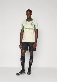 Krämvit sporttröja med gröna detaljer och logotyp, kombinerad med svarta shorts, svarta knästrumpor och svarta fotbollsskor.