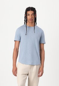 Camiseta de manga corta azul claro, hecha de una tela suave y texturizada. Escote redondo y un corte relajado. Modelo lleva pantalones beige.