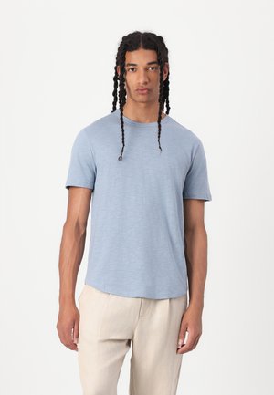 Light blue short-sleeve t-shirt made of a soft, textured fabric. Round neckline and a relaxed fit. Model wearing beige pants.