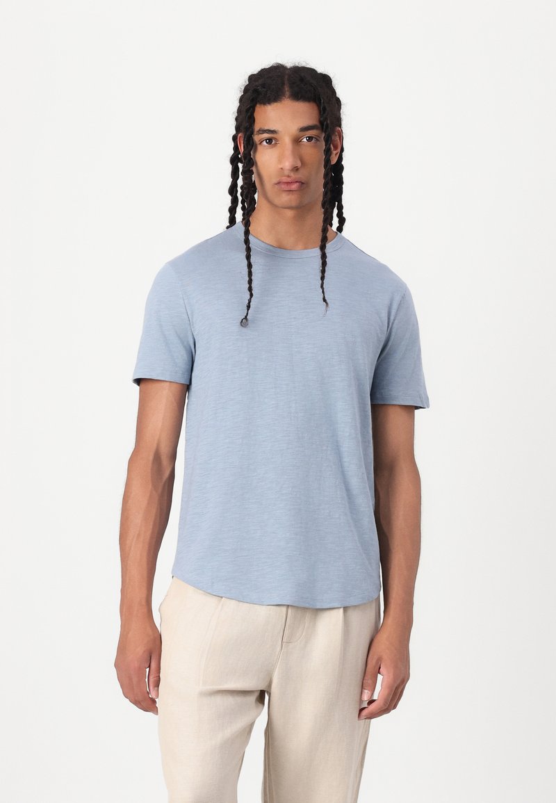 Camiseta de manga corta azul claro, hecha de una tela suave y texturizada. Escote redondo y un corte relajado. Modelo lleva pantalones beige.