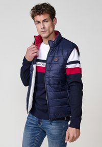 Chaleco acolchado azul marino con forro rojo, cierre de cremallera y parche con logo. Llevado sobre una camisa de manga larga a rayas en azul marino, blanco y rojo, combinada con jeans azules.