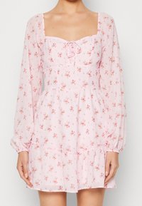 Vestido floral rosa con mangas largas, escote cuadrado, detalle de cordón en el pecho y dobladillo con volantes. Hecho de un tejido liviano y transparente.