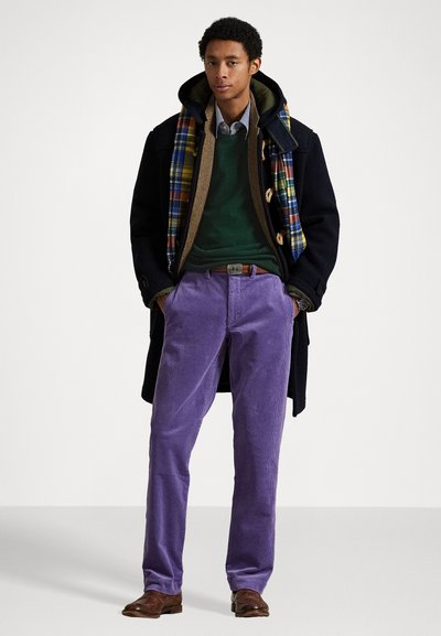Manteau noir sur un pull vert, écharpe écossaise ; chaussures brunes, pantalons en velours côtelé lavande. Des couches et des textures notables dans l'ensemble de la tenue.
