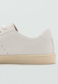 Hellbeiger Sneaker mit glattem Obermaterial, dezenten Perforationen, flachen Schnürsenkeln und einer cremefarbenen Gummisohle. Abgerundete Ferse mit kontrastierenden Nähten.