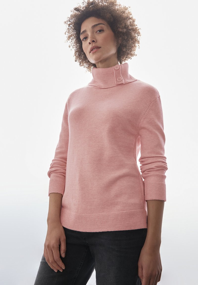 Suéter rosa con un alto cuello vuelto y dos botones. Puños y dobladillo acanalados. Textura suave, diseño ajustado. Llevado con jeans oscuros.