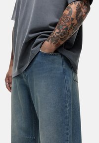 Una maglietta di cotone grigia pende liberamente su pantaloni in denim azzurro chiaro. I pantaloni presentano una finitura sbiadita e cuciture visibili, con le mani nelle tasche.