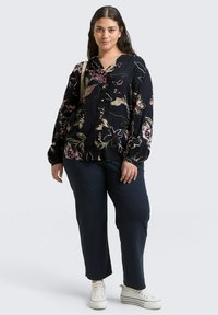 Schwarze Bluse mit floralem Muster, langen Ärmeln und Knopfverschluss, kombiniert mit marineblauen Hosen und weißen Sneakern. Blumen in Pink, Gelb und Grün.