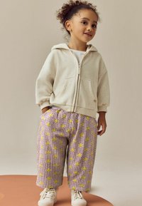 Hellbeige Zip-Up-Hoodie mit einer gesprenkelten Textur, kombiniert mit lila Karo-Hosen, die mit gelben Blumenmustern verziert sind. Weiße Sneaker runden das Outfit ab.