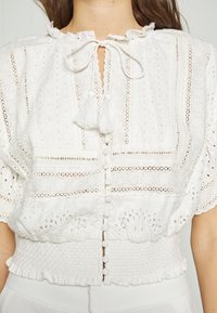 Weiße Bluse mit kunstvoller Stickerei, Lochstickerei-Details, Rüschensamt, Bindebandverschluss und gerafftem Taillenbereich mit Knopfakzenten.