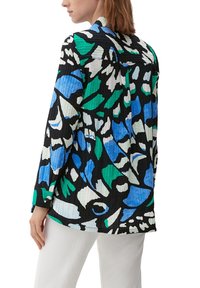 Vrouw gekleed in een langemouwen shirt met een abstract patroon in blauw, groen, wit en zwart, gecombineerd met witte broek, gezien vanaf achter.