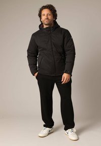 Veste isolante noire avec capuche, dotée de poches zippées sur les côtés et d'une texture lisse, portée avec un pantalon noir et des baskets blanches.