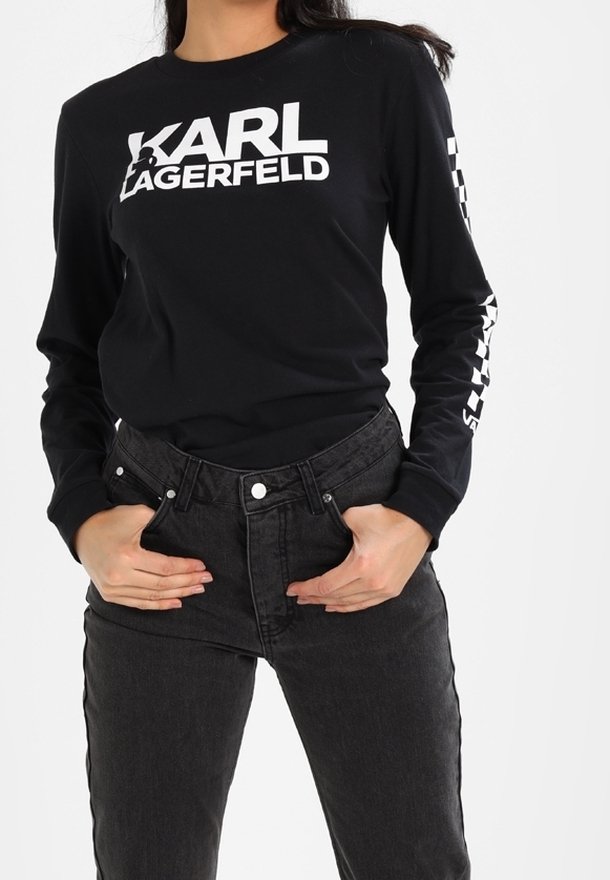 Camiseta negra de manga larga con el texto blanco "KARL LAGERFELD" en el pecho y la manga, combinada con jeans negros de cintura alta. Textura de algodón suave.