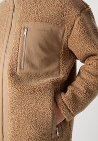 Brun fleece jakke med en blød tekstur, lysbeige stoflomme og en lynlås. Har en opretstående krave og et afslappet fit design.