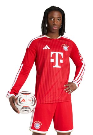 Junger männlicher Fußballspieler im roten FC Bayern München-Trikot hält einen weißen Adidas-Fußball und steht mit ernstem Gesichtsausdruck vor weißem Hintergrund.