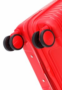 Valise rigide rouge avec surface texturée, quatre roues noires et une fermeture éclair, mettant en avant des accents métalliques sur le côté.