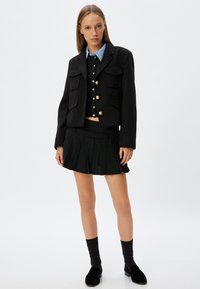Zwarte cropped jas met gouden knopen en zakken, gelaagd over een zwarte overhemdtrui, gecombineerd met een zwarte plissérok. Zwarte sokken en schoenen.