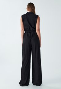 Ženska z dolgimi ravnimi lasmi, oblečena v črn brezrokavnik z širokimi nogami, stoji obrnjena stran od kamere proti enobarvni svetli ozadju.