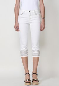 Pantalons capri en denim blanc avec des coutures décoratives sur l'ourlet coupé. Comprend des poches avant et une fermeture à bouton. Porté avec des sandales à plateforme.