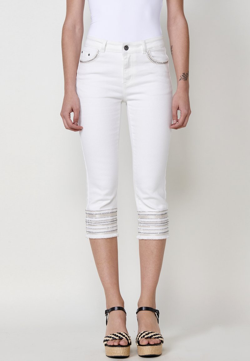 Pantalons capri en denim blanc avec des coutures décoratives sur l'ourlet coupé. Comprend des poches avant et une fermeture à bouton. Porté avec des sandales à plateforme.