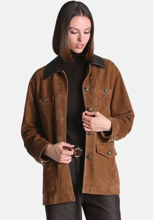 Femme aux cheveux bruns raides portant une veste en daim marron avec des poches boutonnées et un pull à col roulé foncé.