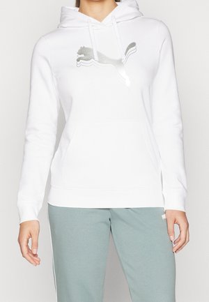 Person trägt einen weißen Hoodie mit silbernem Puma-Logo und hellgrüner Hose, stehend vor einem einfarbigen Hintergrund.