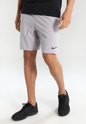 Osoba ubrana w jasnoszare szorty sportowe Nike i czarne buty do biegania Nike, stojąca na tle jednolitego, jasnego tła.