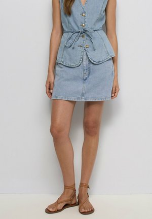 Femme portant une jupe en jean bleu clair et un gilet sans manches assorti avec ceinture à nouer, associée à des sandales plates marron à brides.