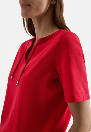 Femme portant un t-shirt rouge vif à manches courtes avec une encolure fendue et des cordons sur un fond clair uni.