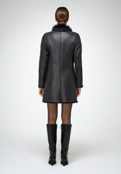Schwarzer Leder-Mantel mit einem Kunstfellkragen, strukturierter Form, taillierter Passform und kontrastierenden Nähten. Kombiniert mit schwarzen kniehohen Stiefeln.