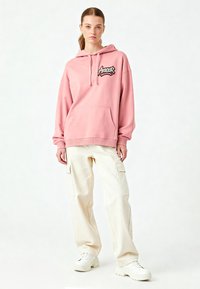 Felpa rosa con logo ricamato "Amour", cappuccio regolabile e tasca frontale. Abbinata a pantaloni cargo beige chiaro e sneaker bianche.